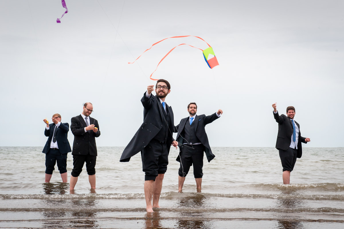 andy and his groomsmen fly kites while standing in the sea in whitstable eknt on his wedding day