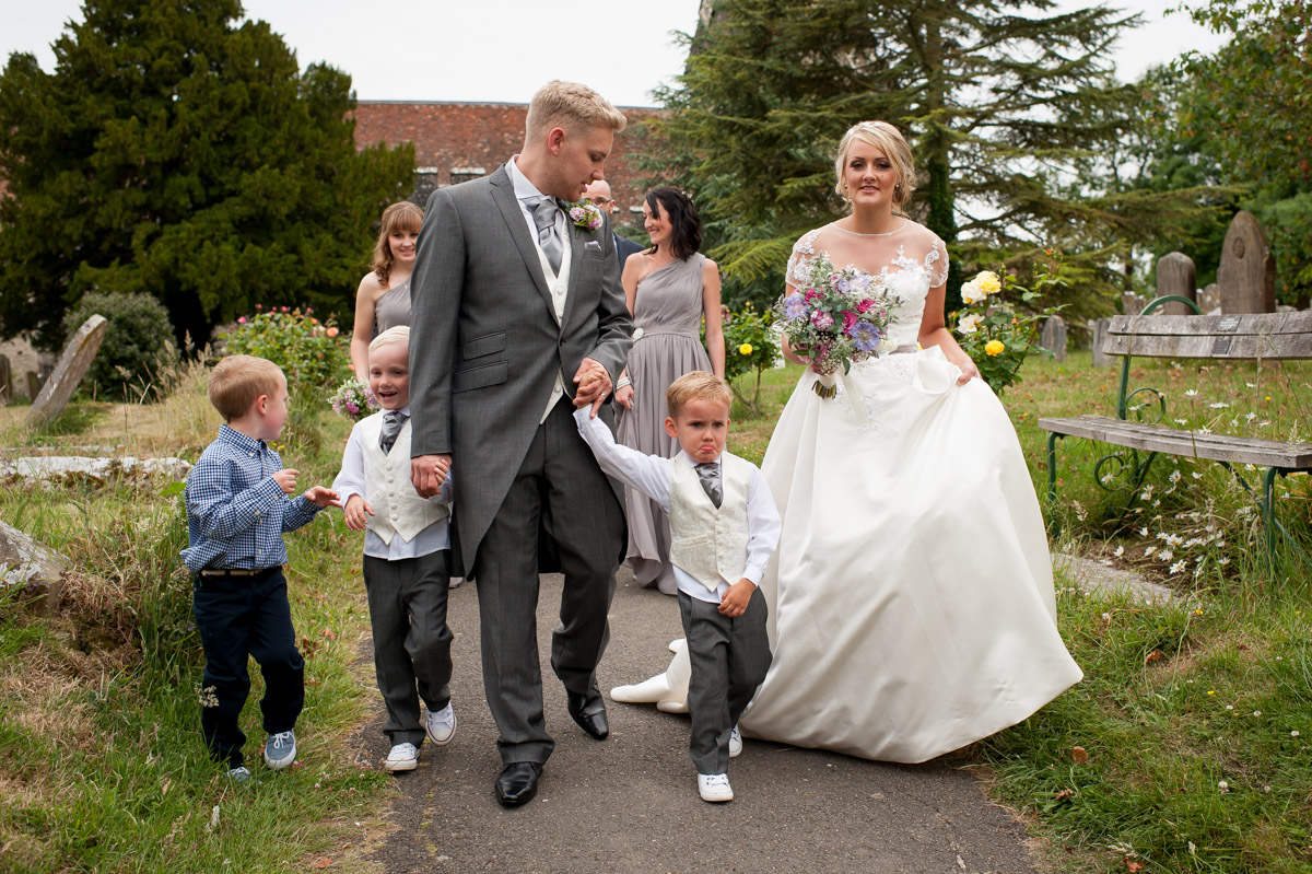 Photo of newly weds and their children walking down church path in wood church, kent
