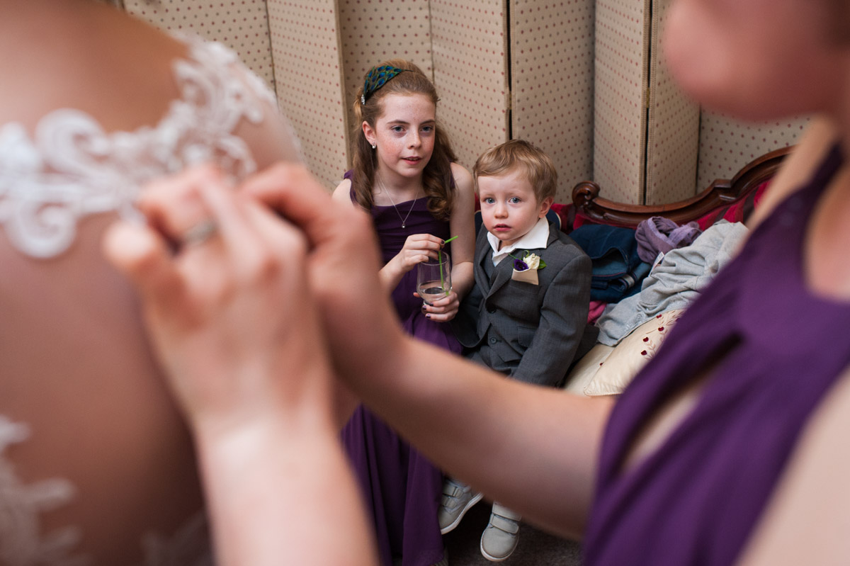Page boy and flower girl watch the brides dress being fastened by the bridesmaid