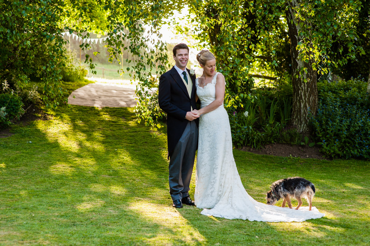 Bride and groom photographed together in the garden at their wedding reception with border terrier dog standing on brides dress