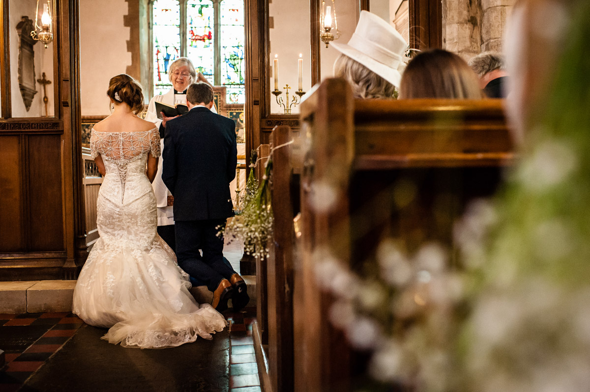 Photograph of Catherine and Jonny kneeling in church while vicar gives blessing on their wedding day