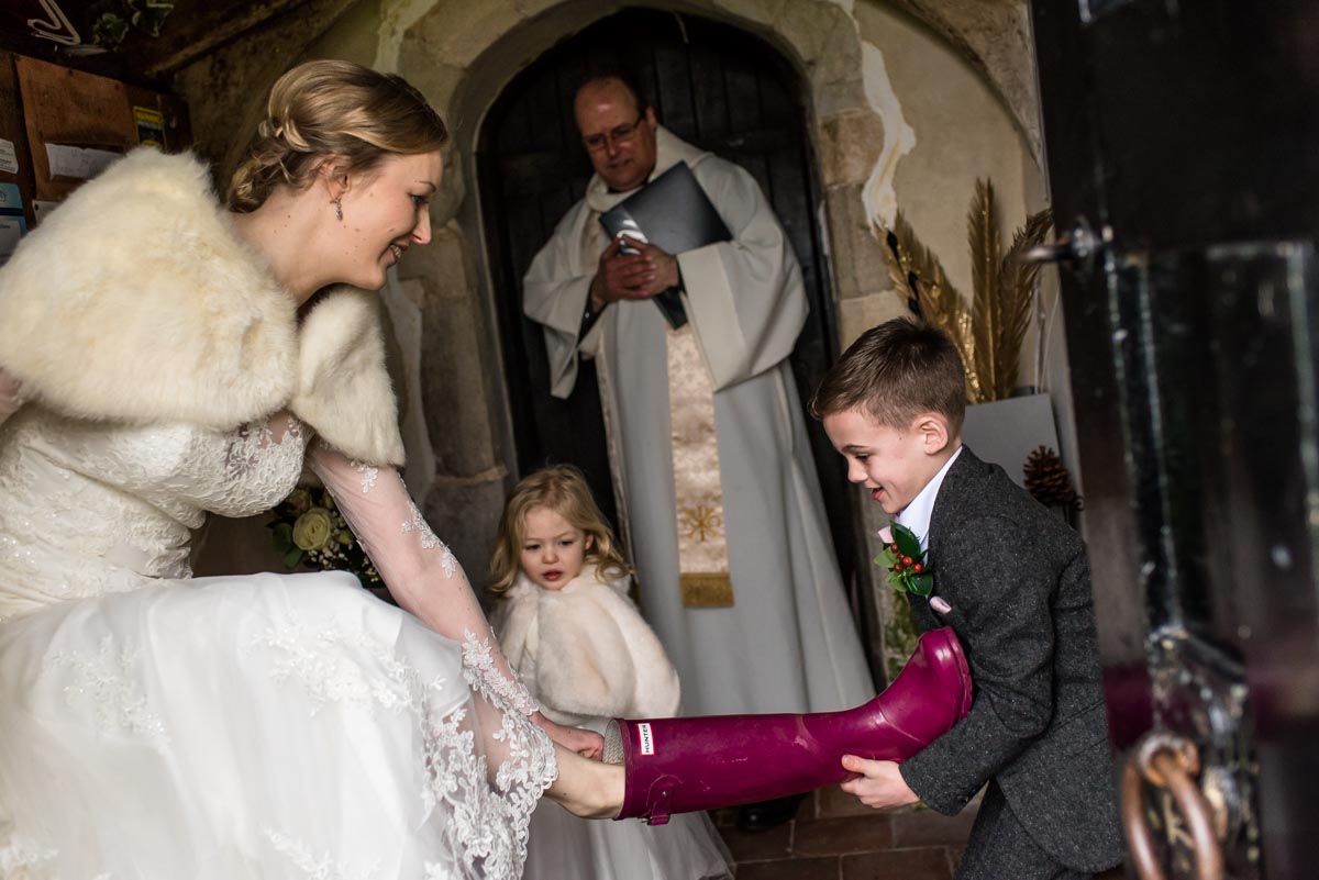 Wedding photography at church. page boy helps bride take off wellington boots