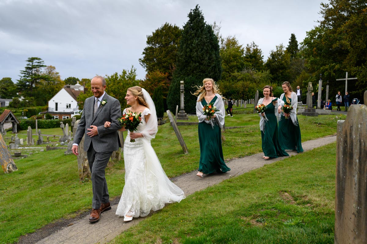 Rebecca, dad and bridesmaids walk path to church on her wedding day